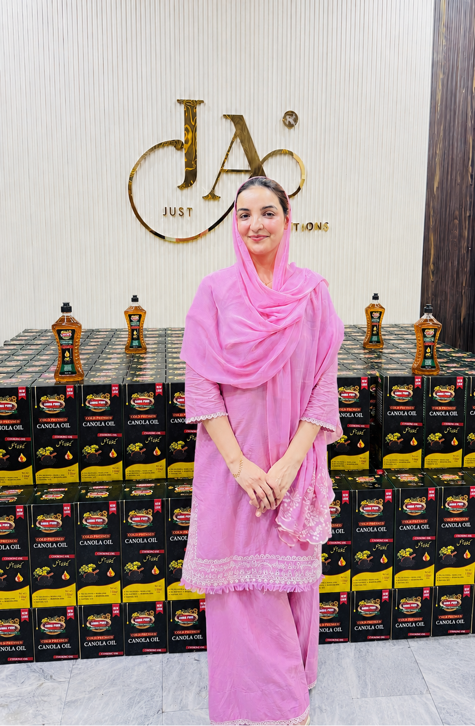 Amina Hussain in Amna Pure Oils Factory wearing a pink traditional outfit and matching headscarf stands in front of a display of stacked black and yellow oil boxes, with a large gold 'JA' Just Amna logo on the wall behind her.