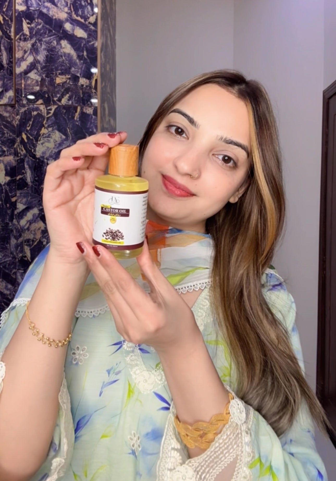 Amina Hussain holding a Castor Oil Plastic Bottle in front of mirror by Just Amna in Summer dress shalwar qameez, dyed hair & wearing gold jewelry