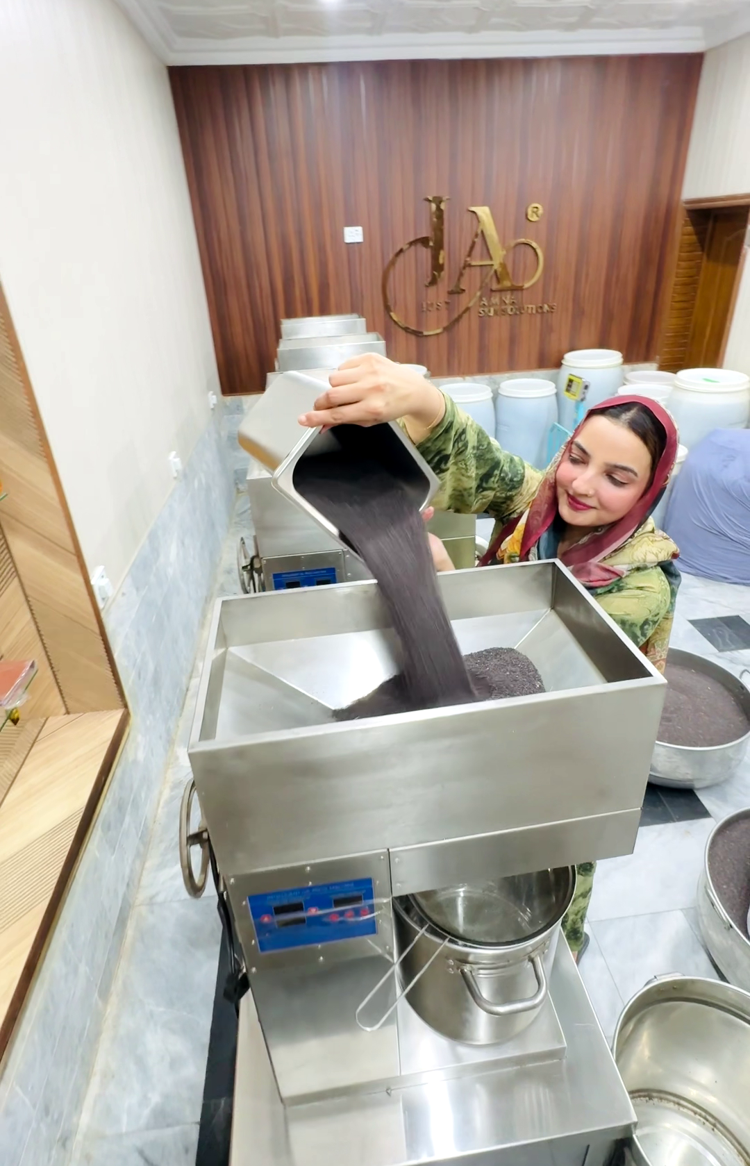 Amina Hussain aka memoona muslima  wearing a green shalwar qameez dupatta and smiles while pouring canola seeds in the cold pressed maching to extract the cooking oil from a white container into a stainless steel machine in Amna Pure oils factory.